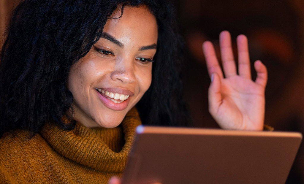 Inclusão digital.  Mulher com cabelo cacheado preto usando um suéter mostarda. Ela está segurando um tablet e levantando uma das mãos.