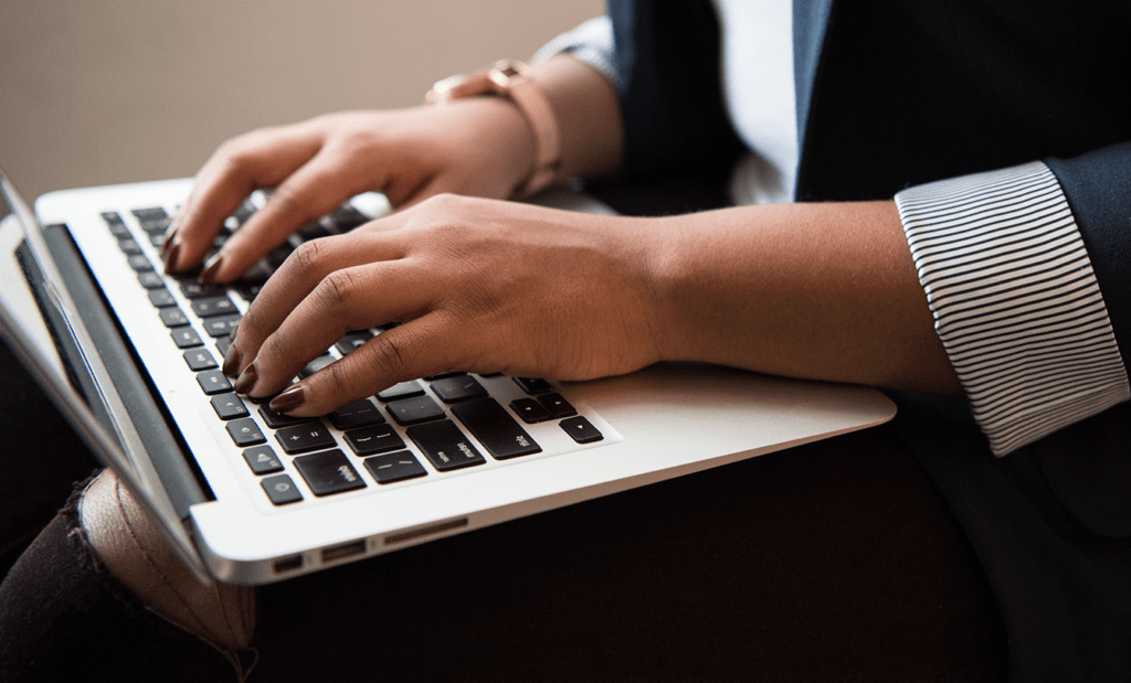 digital. Imagem de uma mão feminina com as unhas pintadas de marrom teclando em um notebook que está por cima da sua perna.