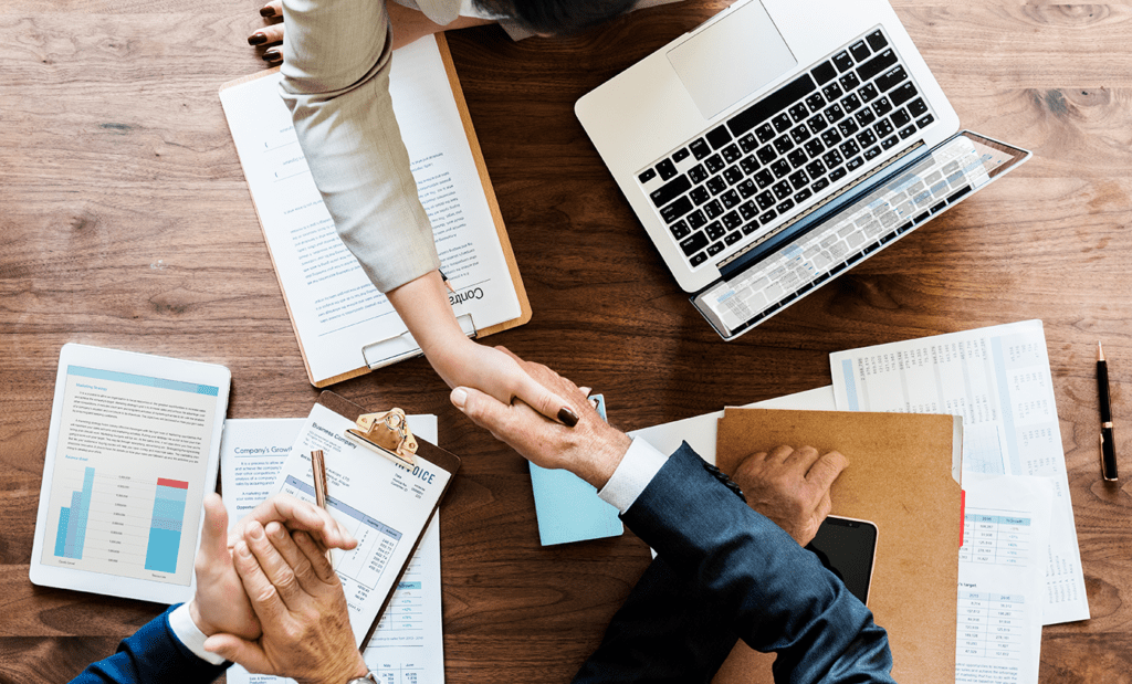 acessível. A imagem de uma mesa de escritório com alguns documentos e notebook. Nela podemos observar uma mão feminina e uma masculina apertando as mãos e ao lado uma mão masculina aplaudindo.