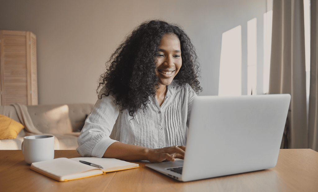acessível. Uma menina negra de blusa social em uma sala usando o notebook em cima de uma mesa e ao seu lado uma xícara branca e um caderno aberto.