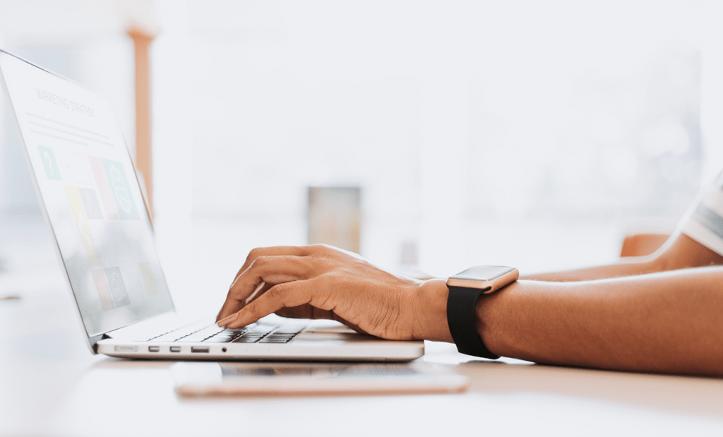 internet. Imagem da mão de um homem branco com relógio no pulso digitando em um notebook que está em cima da mesa.