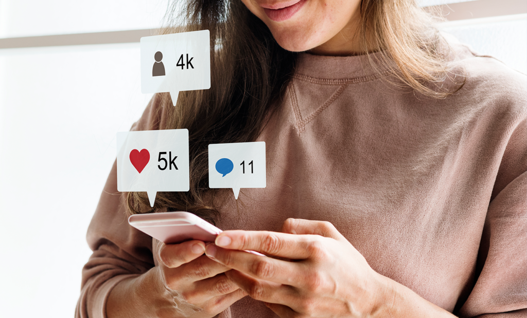 internet. Imagem de uma mulher branca de cabelos longos digitando no celular e nele aparece algumas notifciações de curtidas, comentários e seguidores..