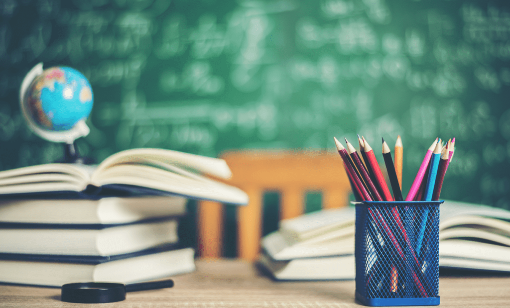 educação. Imagem de uma mesa de estudo com alguns livros empilhados e outros abertos em cima da mesa. Do lado direito podemos observar um porta lápis com alguns lápis dentro e do lado esquerdo uma lupa.