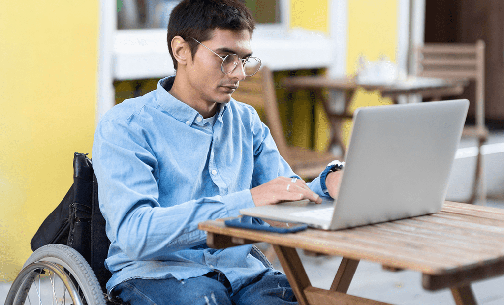 educação. Homem jovem utilizando uma cadeira de rodas enquanto trabalha em um notebook. Ele está sentado em uma mesa de madeira ao ar livre, vestindo uma camisa jeans clara e óculos. Ao fundo, há outras mesas e cadeiras.