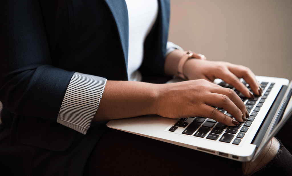 Atualização. Imagem de uma mulher negra com roupa social azul escura, unhas vermelhas e relógio dourado, senta usando o notebook. 