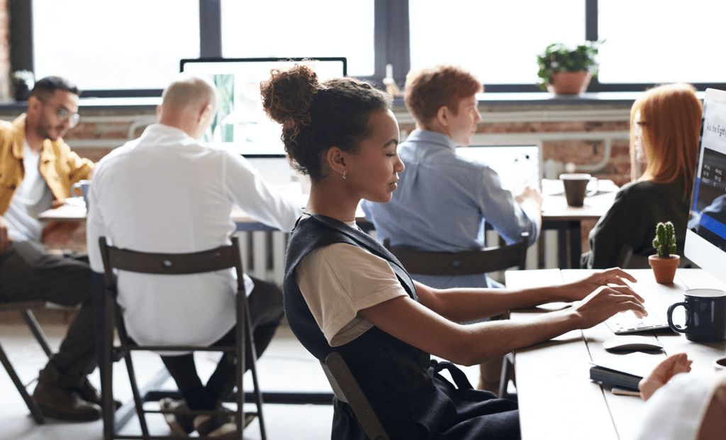 estudo. Imagem de um escritório de trabalho com várias pessoas trabalhando. No foco da imagem aparece uma menina negra de cabelo preso, mexendo no computador. 