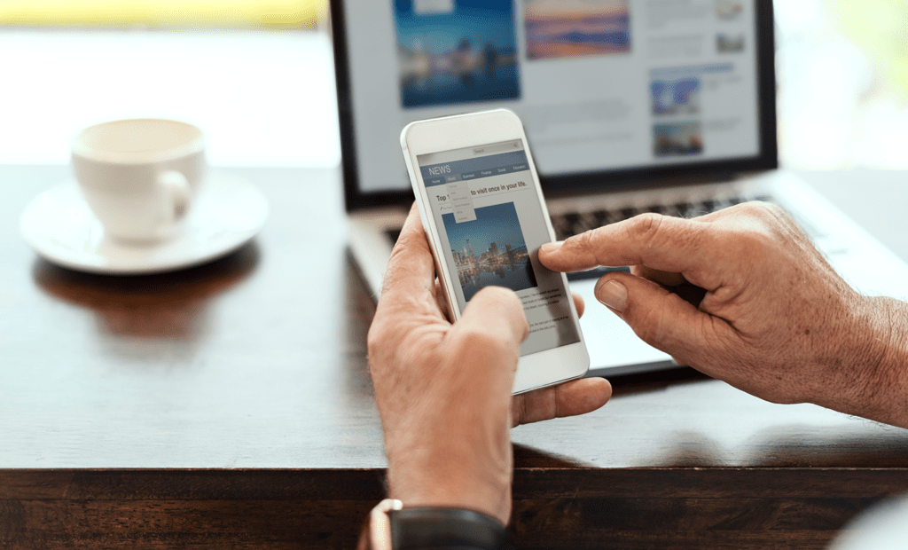 acessível. Imagem de um mão branca, masculina e de aparência idosa mexendo no celular. No fundo da imagem podemos observar um computador aberto em cima de uma mesa com umca xícara de café ao lado. 