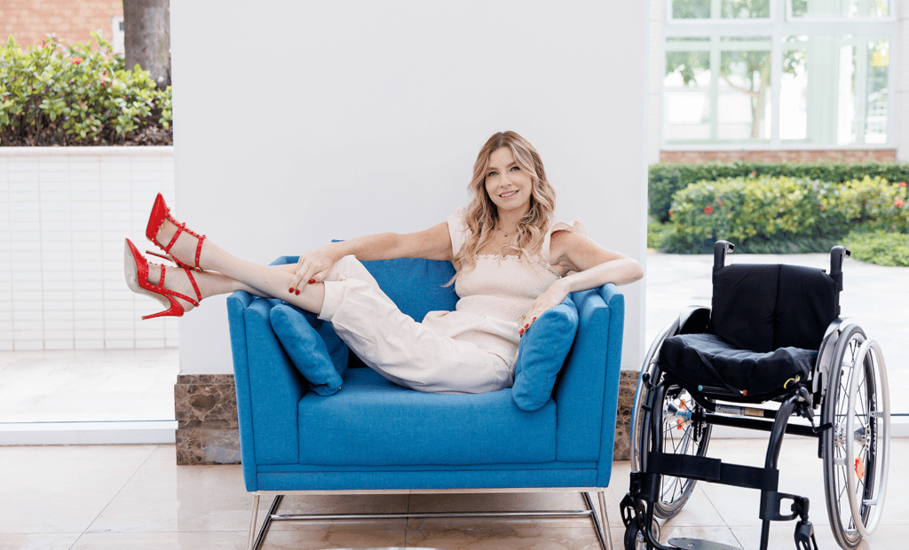marketing. Foto na nossa embaixadora Andrea sentada em um poltrona com as pernas para o ar, ao lado da poltrona está sua cadeira de roda. Andrea veste um conjuto na cor bege e usa um salto alto na cor vermelha. 
