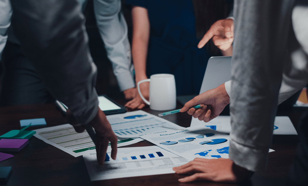 DEI. Imagem de uma mesa de reunião com alguns papeis por cima, nela também podemos observar pessoas com roupas sociais apontando para a mesa como se estivessem em uma reunião.