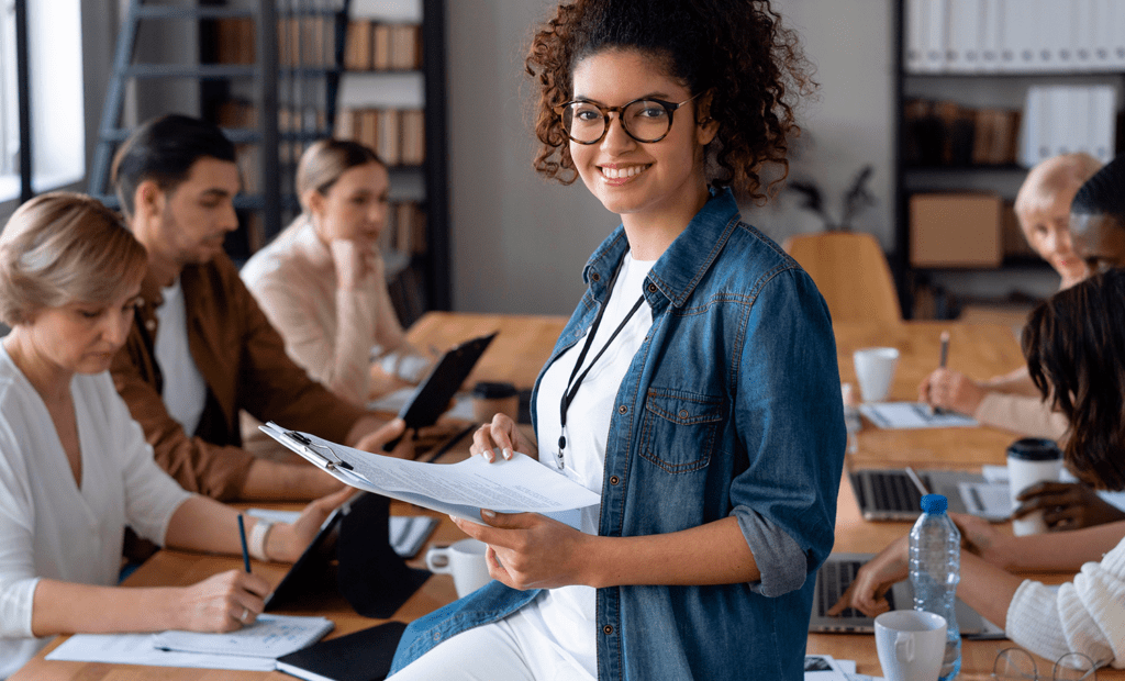 DEI. Imagem de uma biblioteca com algumas pessoas estudando e no centro da imagem em foco temos uma menina negra de pele pouco retinta, usando óculos e sorrindo, segurando uma prancheta.