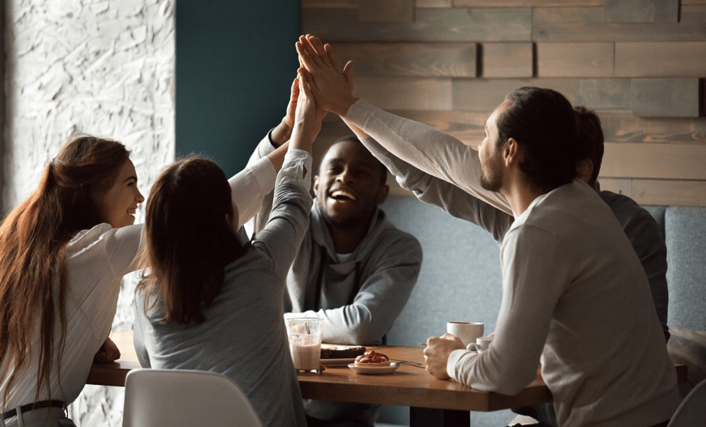 Diversidade. Na imagem podemos observar um grupo s de pessoas sentada em um café, todos eles estão com as mãos para alta bantendo com as mãos um dos outros. 