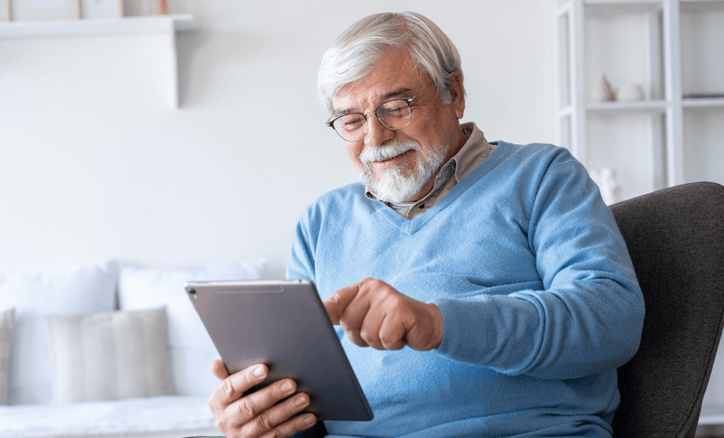 deficiência. Imagem de um senhor de idade branco e de cabelos brancos, usando uma casaco azul. Ele está usando um tablet e sorrindo sentado na sala.
