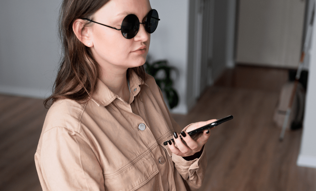 deficiência. Imagem de cega usando o celular. Ela está com uma blusa bege e usa óculos escuros.