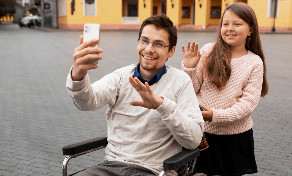 entretenimento. Imagem de duas pessoas na rua sorrindo para um foto, uma delas é um homem branco, cadeirante que segura o celular e tira foto e atrás dele temos uma crianças, meninas, brancas com cabelos longos acenando para foto.