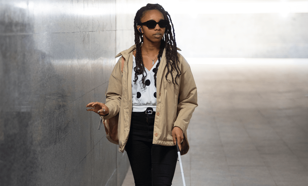 entretenimento. Imagem de uma mulher negra deficiente visual, com tranças e óculos escuros e bengala andando por um corredor.