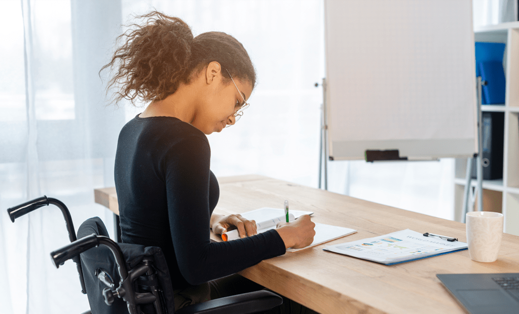 Conscientização. Imagem de uma menina negra usando óculos e blusa preta de manga longa, cadeirante, estudando em uma sala de reunião.
