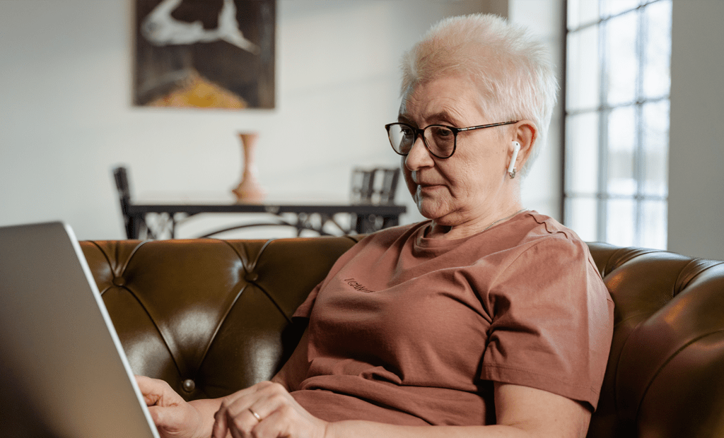 Conscientização. Imagem de uma senhora de idade branca, sentada no sofá mexendo no notebook. A senhora tem cabelo branco e usa óculos de grau.