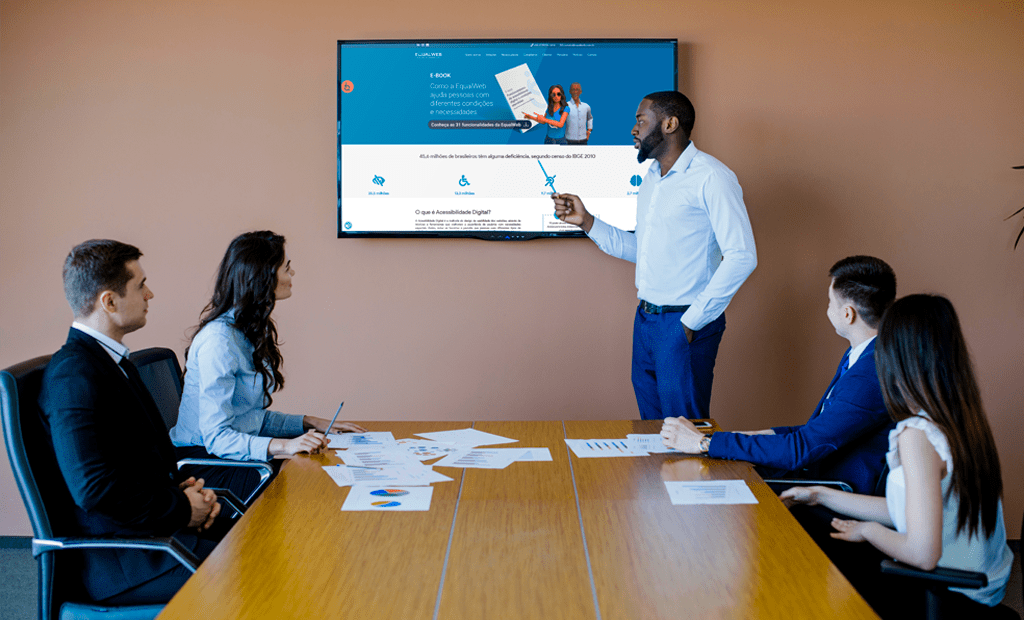 Design acessível. Imagem de uma sala de reunião com algumas pessoas e uma delas esta apresentando o site da Equalweb para o restante do grupo.