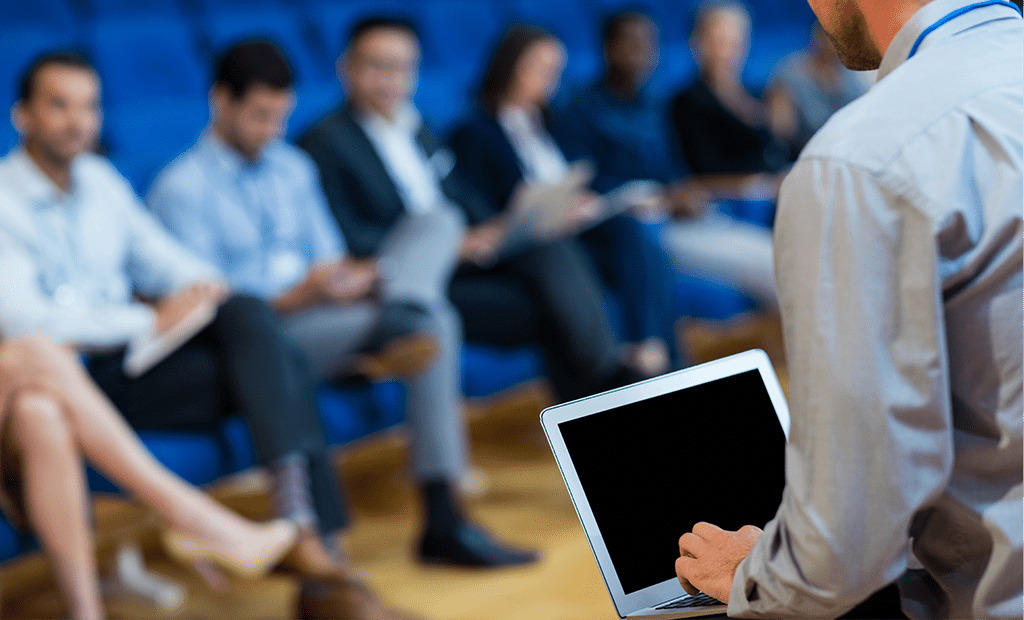 Evento. Imagem de uma plateia com alguns convidados. A imagem apresenta um leve embaçado no público e no foco central, temos um homem de roupa social, branco em um pubito mexendo no computador.