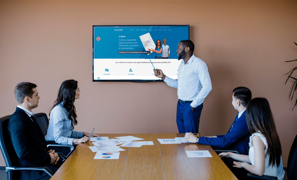 Evento. Imagem de um grupo de pessoas em uma sala de reunião e na tela da sala o site da EqualWeb aberto sendo apresentado. 