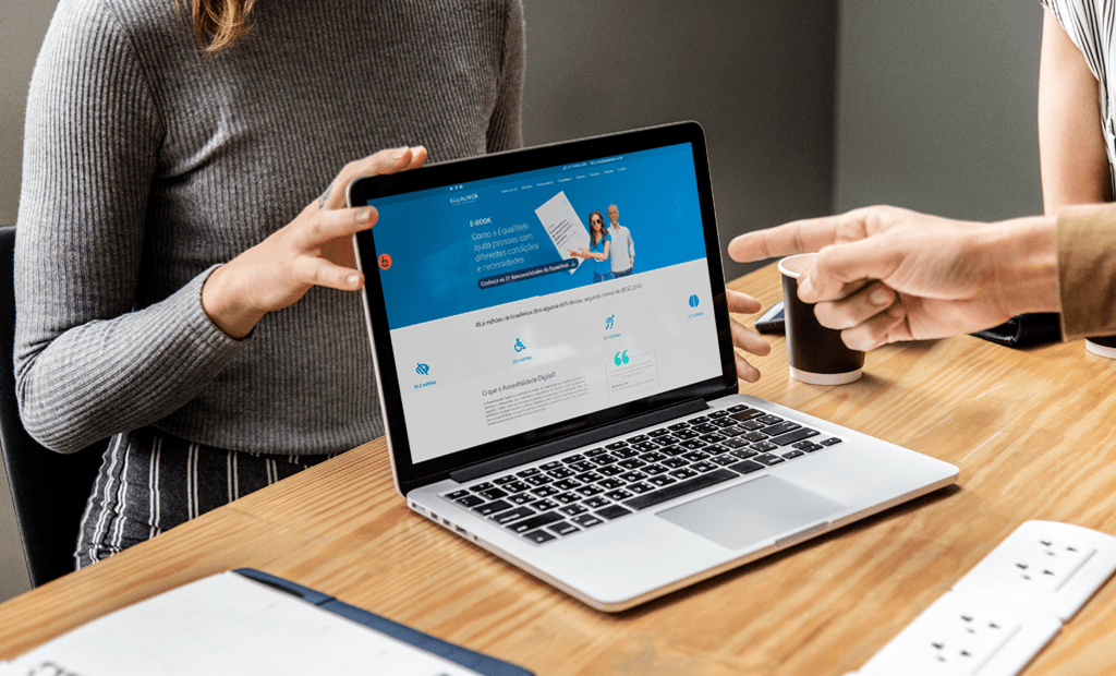 DEIA. Imagem de um notebook aberto em cima de uma mesa com a página do site da EqualWeb aberta. Em volta da mesa aparecem algumas mãos de pessoas apontando para o site. 