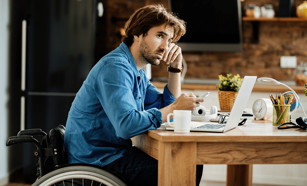 acessibilidade digital. Imagem de um homem branco, cabelos longos, cadeirante, sentado em uma mesa de madeira e mexendo no notebook. 