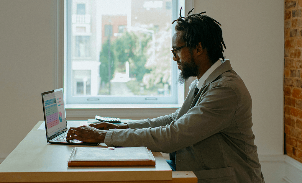 Tedências. Imagem de um homem negro, vestido rouupa social, de óculos e barba. Mexendo no computador. 
