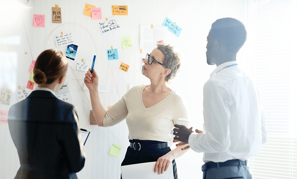 Comunicação: Imagem de uma sala de reunião com alguns papéis e escritos em um quadro em branco. Na frente do quadro, temos duas mulheres brancas e um homem negro, todos vestindo roupas sociais.