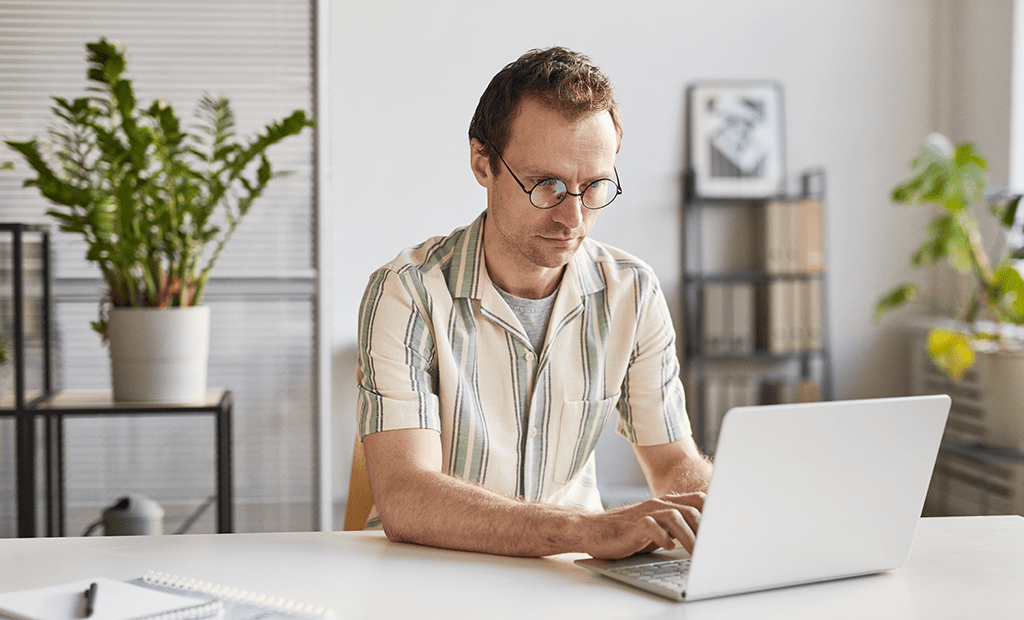 Acessíveis. Imagem de um homem branco e de óculos com cabelos ruivos, mexendo no notebook em uma sala.