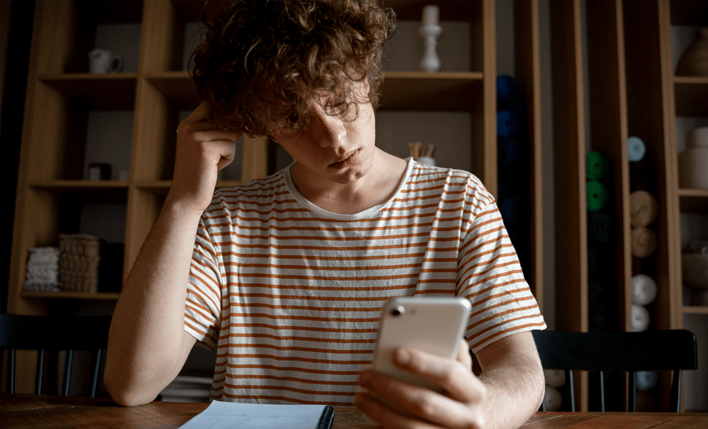 Libras. Jovem olhando para o celular enquanto coça a cabeça, sentado à mesa com caderno e caneta, em ambiente interno com prateleiras ao fundo.
