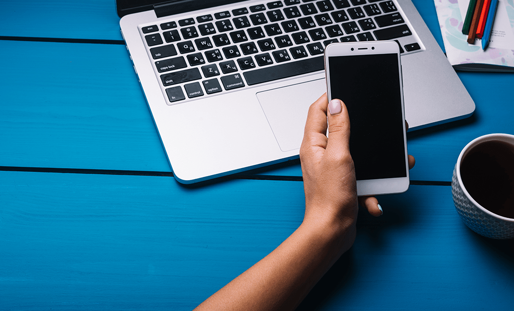 Responsividade. Imagem de uma mão feminina segurando um celular e a sua frente um notebook aberto.