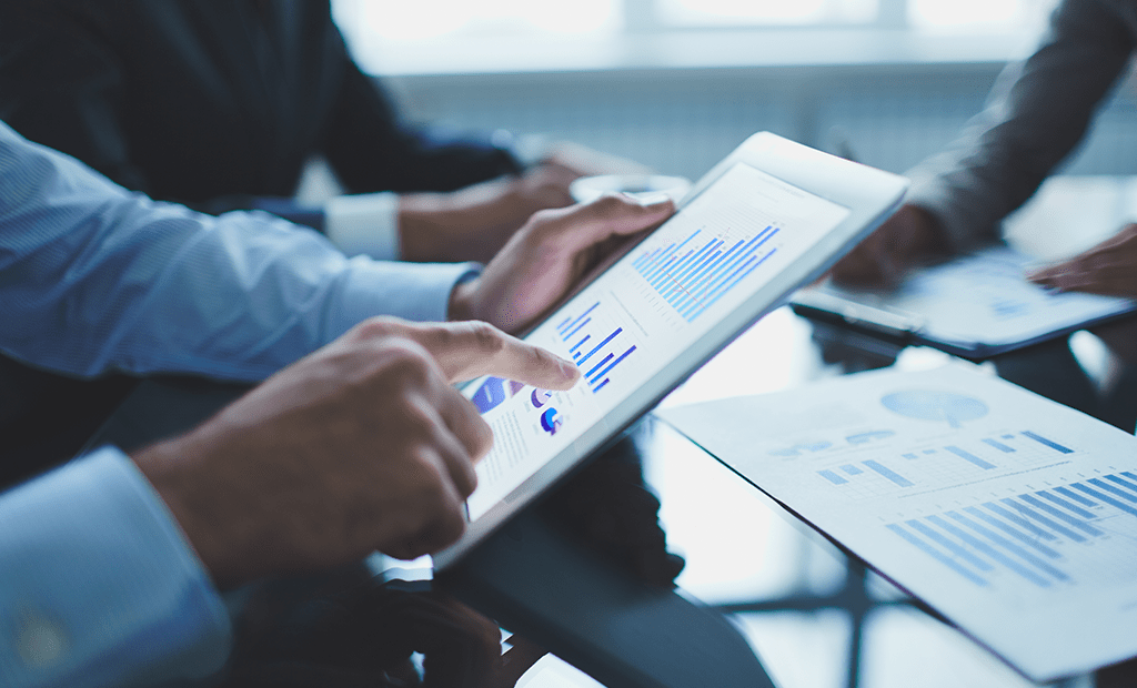 ROI. Pessoas estão analisando gráficos e dados em um tablet. O tablet exibe gráficos de barras e outros elementos visuais, enquanto há documentos com gráficos semelhantes sobre uma mesa de vidro.
