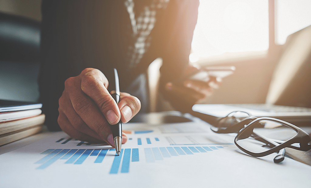 ROI. Um profissional segura uma caneta enquanto marca ou analisa dados representados em gráficos de barras impressos em papel. Sobre a mesa estão óculos e outros itens de trabalho, como documentos e um laptop. 