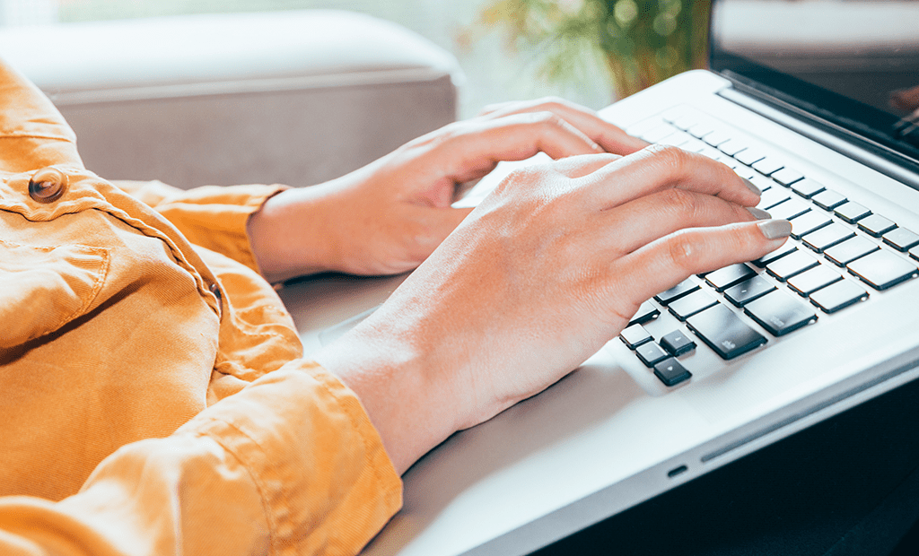 ABNT. Mulher sentada em um sofá digitando em um notebook. Ela veste uma blusa amarela.
