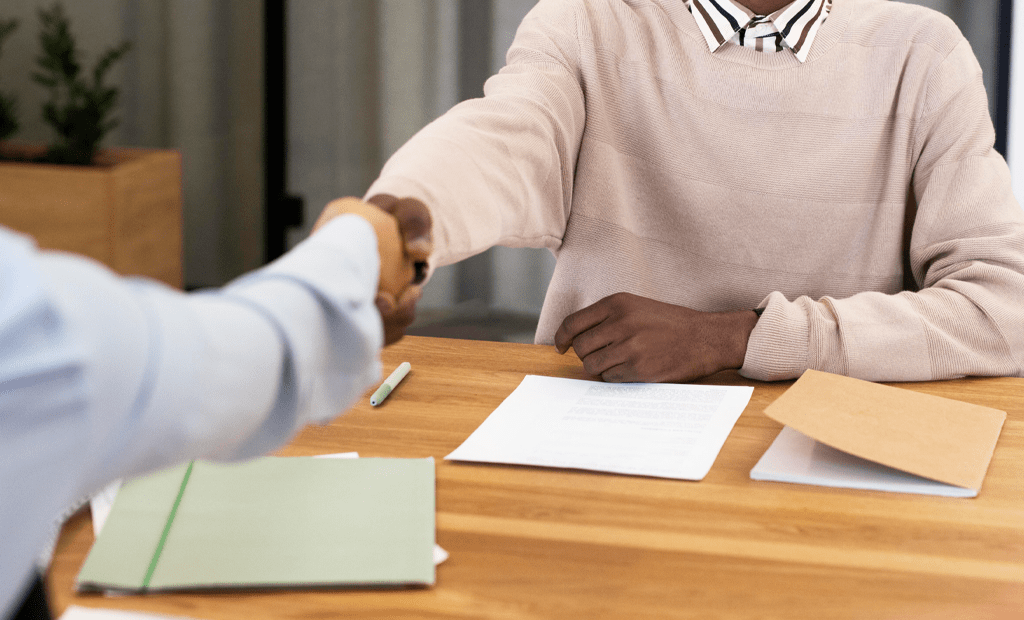 ABNT. duas pessoas apertando as mãos sobre uma mesa de madeira. Na mesa, há documentos, uma pasta verde, um caderno marrom e uma caneta. Uma das pessoas veste um suéter bege sobre uma camisa listrada, enquanto a outra usa uma camisa azul clara.