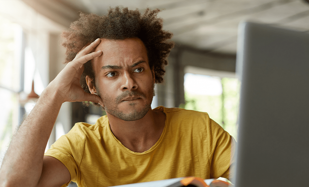 Leitores de tela. Homem jovem sentado em frente a um computador, com uma mão apoiada na cabeça. Ele veste uma camiseta amarela e está em um ambiente interno.