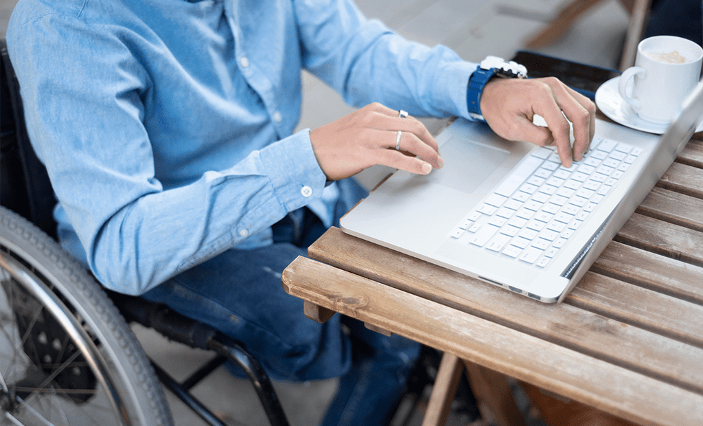NBR-17225. Homem usando cadeira de rodas, vestindo camisa jeans azul, digitando em um notebook prateado sobre uma mesa de madeira. Ao lado do notebook, há uma xícara branca com café da.