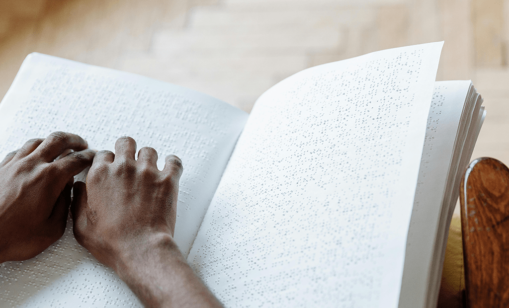 NBR-17225. Mãos de uma pessoa negra lendo um livro em braille. O livro está aberto sobre o colo.