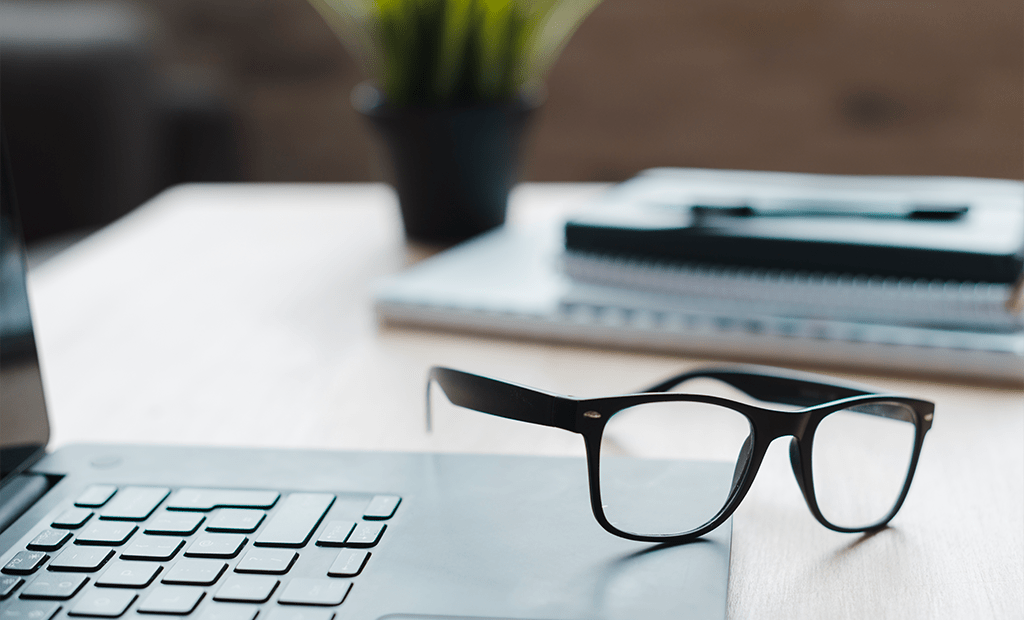 NBR-17225. Óculos de grau com armação preta apoiados sobre um notebook aberto, em uma mesa de madeira clara. Ao fundo, há cadernos empilhados e uma planta decorativa em um vaso.