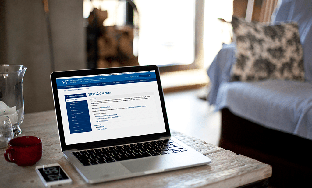 WCAG. Notebook em uma mesa exibindo a página 'WCAG 2 Overview' do site da W3C. Ao lado, uma xícara vermelha e um celular. Ambiente doméstico ao fundo.