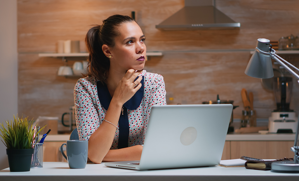 WCAG. Mulher pensativa diante de um notebook em uma cozinha organizada, com caneca, caderno e luminária na mesa.