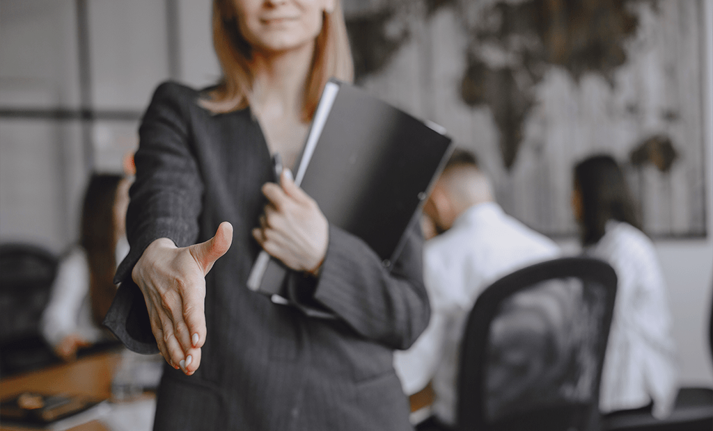 wcag. Mulher oferecendo aperto de mão, segurando pasta preta, com pessoas reunidas ao fundo em escritório
