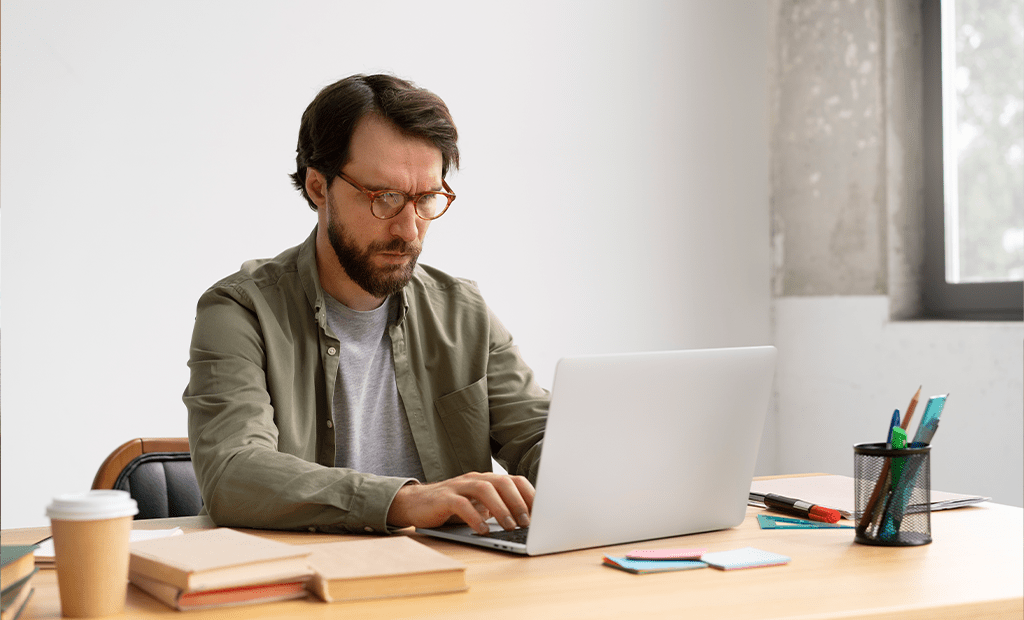 wcag. Homem com óculos trabalhando concentrado em laptop, sentado à mesa com livros, café e materiais de papelaria