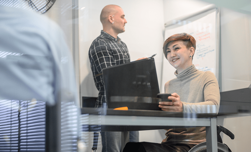 LBI. Mulher cadeirante trabalhando em escritório com notebook e celular, enquanto colega escreve em quadro branco.