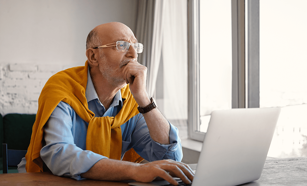 LBI. Homem idoso usando notebook, com expressão pensativa, sentado próximo à janela.