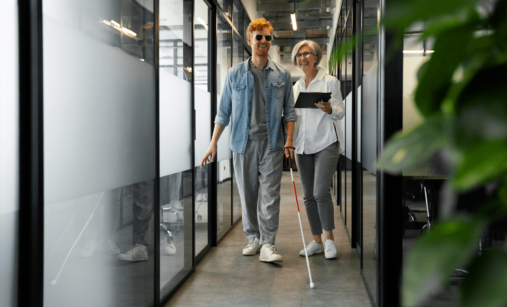 LBI. Homem com deficiência visual caminhando por corredor corporativo com bengala longa, acompanhado por colega segurando um tablet.