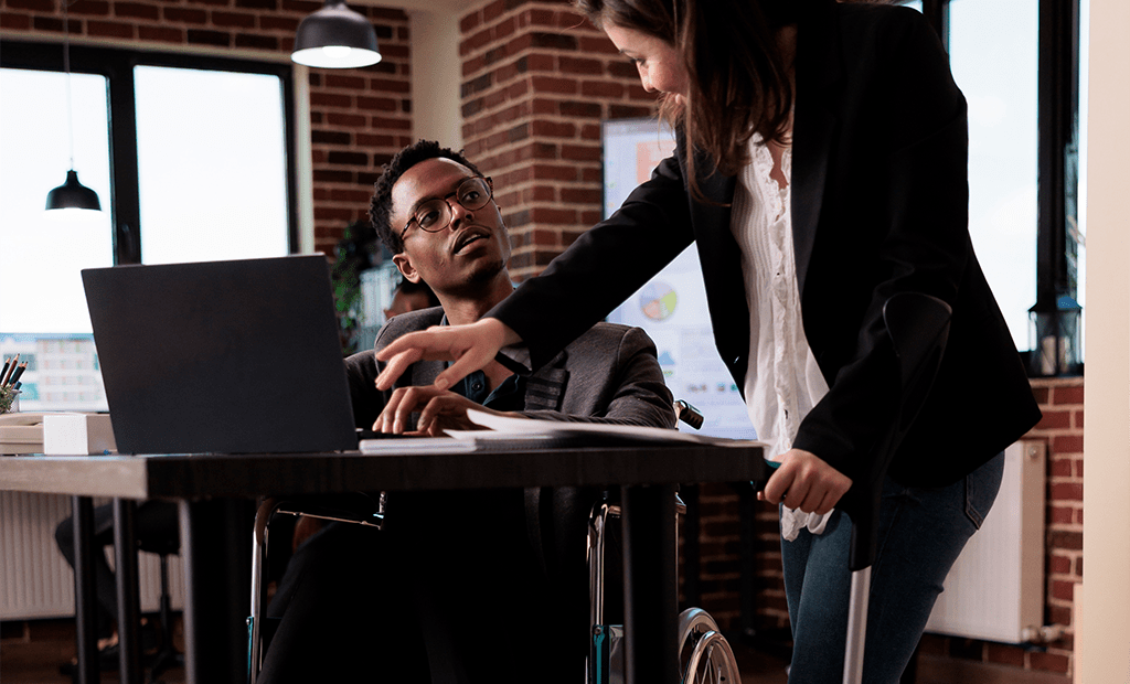 LBI. Homem cadeirante trabalhando em frente ao notebook, enquanto colega com muleta aponta para documentos na mesa.