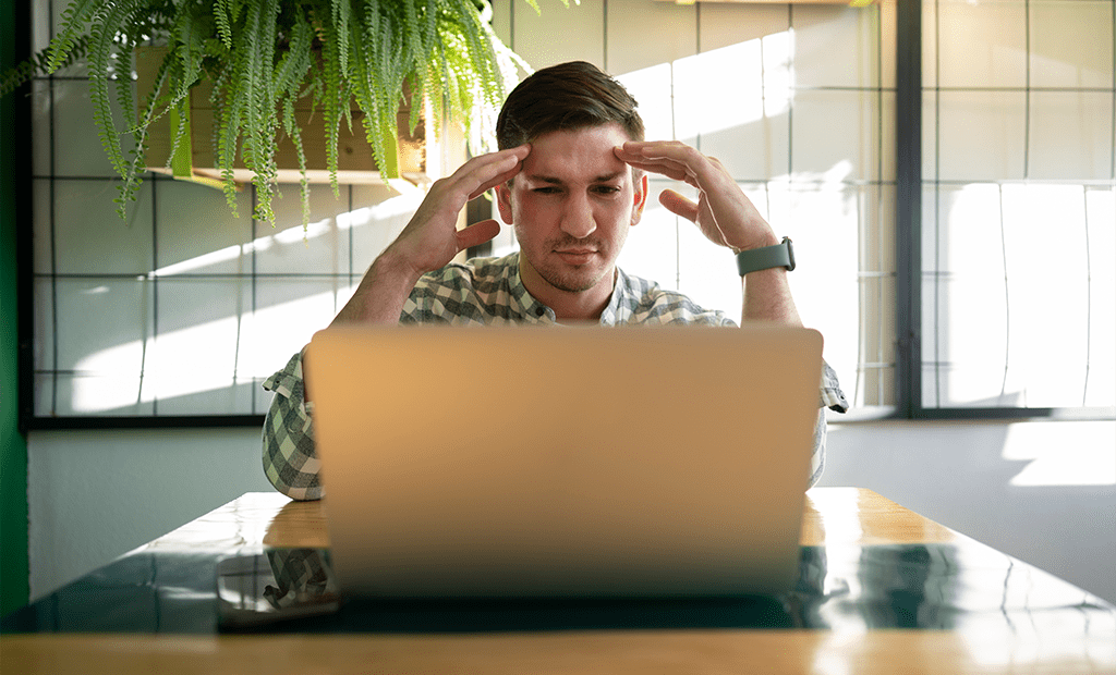 Autismo. Homem com expressão de frustração olha para notebook, sentado em mesa com celular ao lado e plantas ao fundo, em ambiente iluminado.