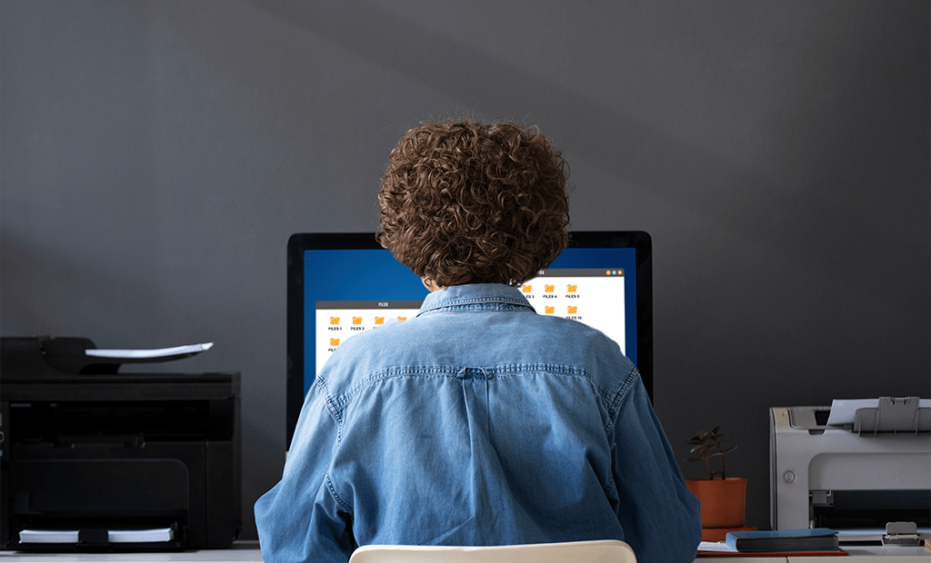 Autismo. Pessoa com cabelo cacheado e camisa jeans usando computador de mesa, vista de costas, em ambiente de escritório com impressoras ao redor.