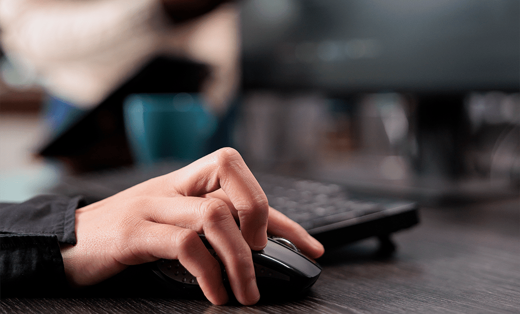 Autismo. Mão usando mouse sobre mesa escura, com teclado e monitor ao fundo em ambiente de trabalho moderno.